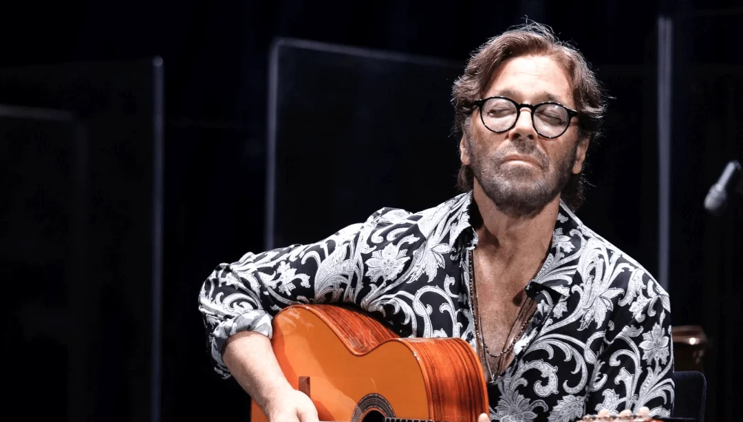Al Di Meola, con gafas redondas y camisa estampada en blanco y negro, tocando su guitarra acústica de color naranja sobre un escenario oscuro