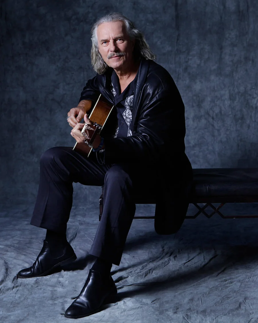 Hans Theessink sentado en un banco con su guitarra acústica sobre fondo oscuro